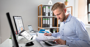 Person sat at desk using calculator, and various screens, using accounts payable automation software