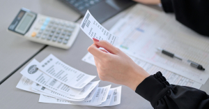 Person going through a pile of paper receipts and inputting data on a computer. Calculator and pen on desk.